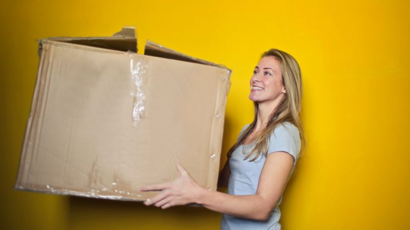 A woman carrying a large cardboard box against a vibrant yellow background, smiling. — Finding out how to Get 6-pack Abs