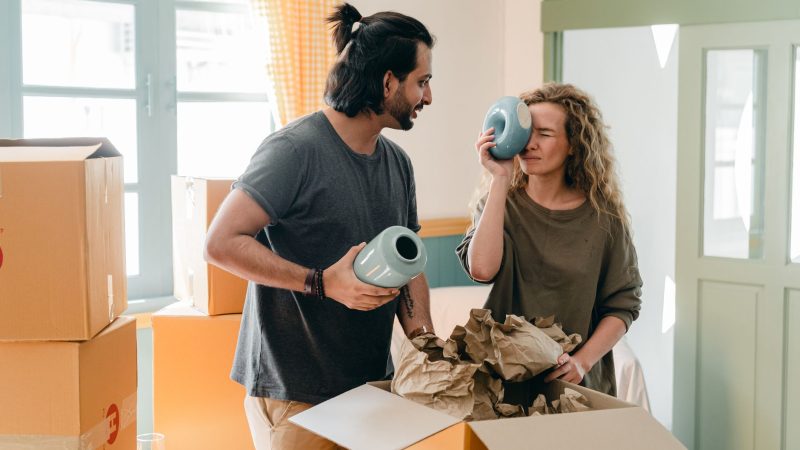 Cheerful ethnic man in smart watch with blue ceramic vase interacting with funny girlfriend with closed eyes and big souvenir